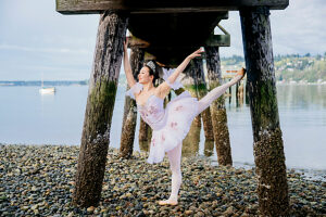 Ballerina poses gracefully on rocky shore beneath a wooden pier extending over calm water. WA, USA