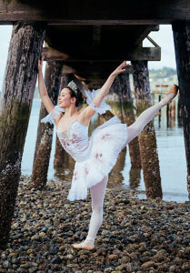 Ballerina in white tutu poses gracefully on pebbled beach under pier beams. WA, USA
