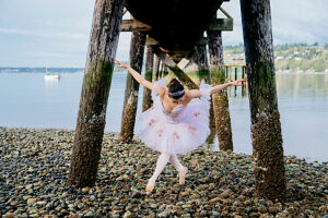 Ballerina in white tutu poses gracefully under a seaside wooden pier on a rocky shore. WA, USA
