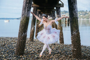 Ballerina dancing on rocky shore under pier with a serene sea and hills in the background.  WA, USA