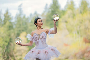 Ballerina wearing a tiara performs with crystal balls in a forest setting. WA, USA