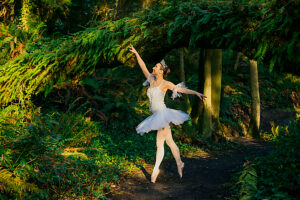 Ballerina in white tutu dancing gracefully in a lush green forest under a moss-covered branch. WA, USA