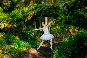Ballerina dances gracefully in a lush green forest under a curved fern-covered archway. WA, USA