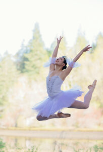 Ballet dancer in mid-air leap against a blurred forest background. WA, USA