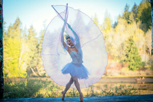 Ballet dancer in white tutu poses gracefully outdoors with sheer fabric, forest backdrop. WA, USA