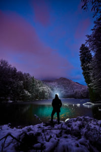 Evening mood and the first snow at the castle pond at Hohenburg near Lenggries, man stands at the lake in the evening dusk with forehead lamp