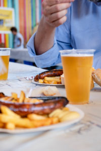A man in a blue shirt is sitting in front of a plate of bratwurst and fries, and beer from plastic cups A man in a blue shirt is sitting in front of a plate of bratwurst and fries, and beer from plastic cups