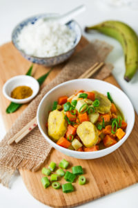 Asiatisches Curry mit Kochbanane und einer Schale weißem Reis auf Holzbrett mit Stäbchen und Deko Asiatisches Curry mit Kochbanane und einer Schale weißem Reis auf Holzbrett mit Stäbchen und Deko