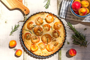 Aprikosen-Schmand-Tarte auf Gartentisch mit Aprikosen Rosmarin Verzierung, Schale mit Aprikosen und Schneidebrett angeschnitten am Bildrand. Aprikosen-Schmand-Tarte auf Gartentisch mit Aprikosen Rosmarin Verzierung, Schale mit Aprikosen und Schneidebrett angeschnitten am Bildrand.