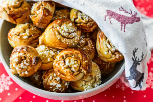 Zimtschnecken in einer Schüssel, halb bedeckt von einem Tuch mit Rentieren auf einer roten Weihnachtsdecke Zimtschnecken in einer Schüssel, halb bedeckt von einem Tuch mit Rentieren auf einer roten Weihnachtsdecke
