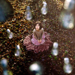 Woman with serious expression sitting on forest floor,surrounded by light bulbs -drops Woman with serious expression sitting on forest floor,surrounded by light bulbs -drops