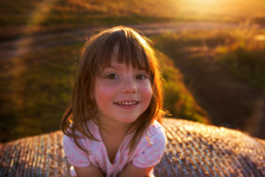 4-6 years old girl sitting on hay bale, smiling in the camera, evening light 4-6 years old girl sitting on hay bale, smiling in the camera, evening light