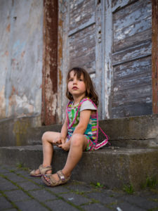 4-6 years old child with coloured dress sitting alone on stairs in front of old wooden door 4-6 years old child with coloured dress sitting alone on stairs in front of old wooden door