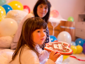 4-6 jähriges Mädchen knabbert in ihren Zimmer voller Luftballons an einem Lebkuchenherz, Mutter im Hintergrund 4-6 jähriges Mädchen knabbert in ihren Zimmer voller Luftballons an einem Lebkuchenherz, Mutter im Hintergrund