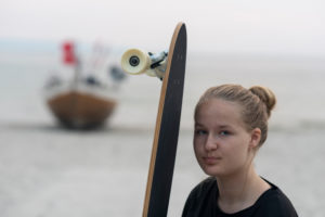Mädchen mit Longboard steht vor einem Fischerboot Mädchen mit Longboard steht vor einem Fischerboot
