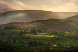 Sunlight falling upon green fields with trees and hedgerows with mountains in the background in England 