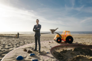 Businessman figurine standing on sand with toy digger, investment in home ownership