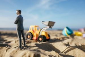 Businessman figurine standing on sand with toy digger, investment in home ownership