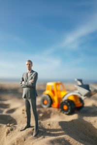 Businessman figurine standing on sand with toy digger, investment in home ownership