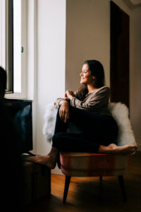 Smiling young woman looking through window while sitting on chair at home Smiling young woman looking through window while sitting on chair at home