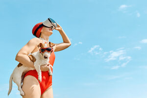 Woman in red swimsuit with VR glasses holding a Jack Russell Terrier wearing sunglasses outdoors