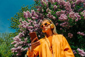Stylish woman in yellow shirt listening to music with headphones near flowering lilac tree outdoors