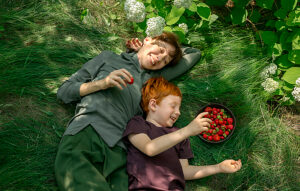 Cheerful mother and son with red hair eating strawberries on grass outdoors