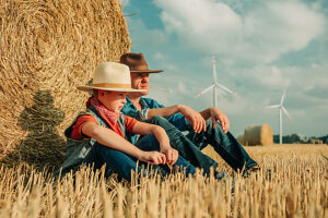 Father and son in cowboy hats relaxing by hay bale in wheat field
