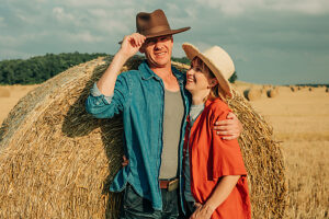 Couple in hats embracing near hay bale in summer wheat field