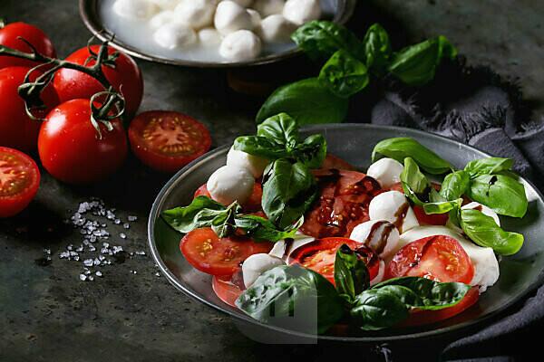 Hand with a white pepper shaker seasoning a fresh salad bowl mixed green  leaves, eggs, black olives and tomato on a wooden table with cutlery.  Blurred nature background. Vertical photography Stock Photo