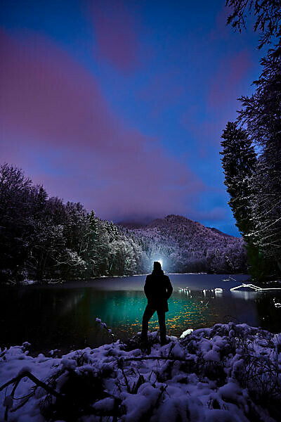 Evening mood and the first snow at the castle pond at Hohenburg near Lenggries, man stands at the lake in the evening dusk with forehead lamp